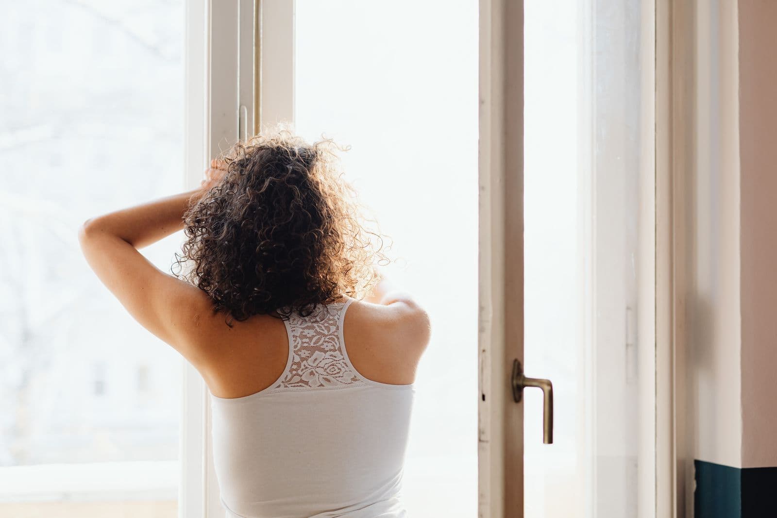 A woman seen from behind stands by a bright window with her hands raised to her head in soft morning light.