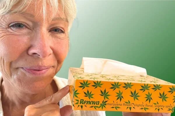 Emotions therapist Becca Williams smiling as she points to a yellow Renova tissue box decorated with green cannabis leaf patterns, containing facial tissues labeled "Marijuana Flower," highlighting a conscious cannabis shift in everyday packaging