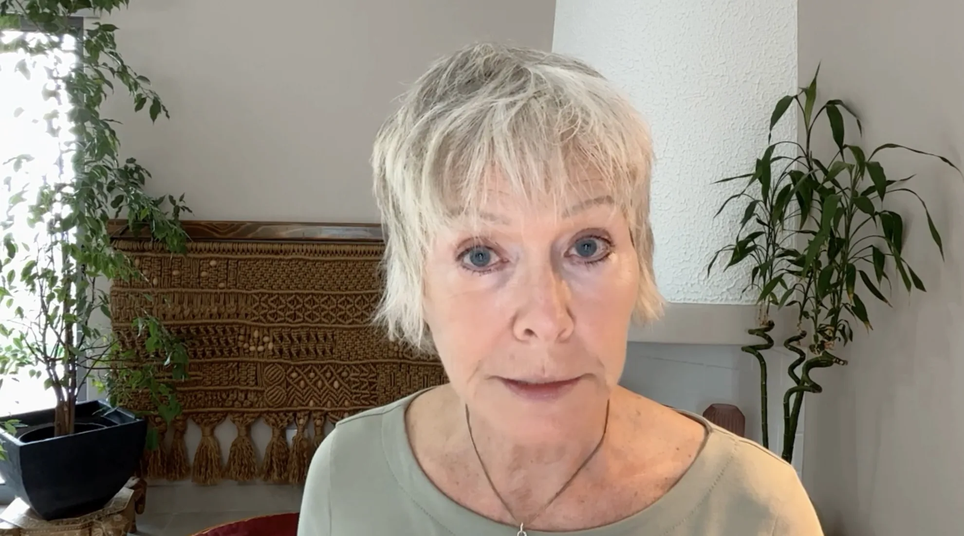 A close-up portrait of Becca Williams wearing a light green top while discussing collective trauma and stress, set against a background of houseplants and woven wall decor.